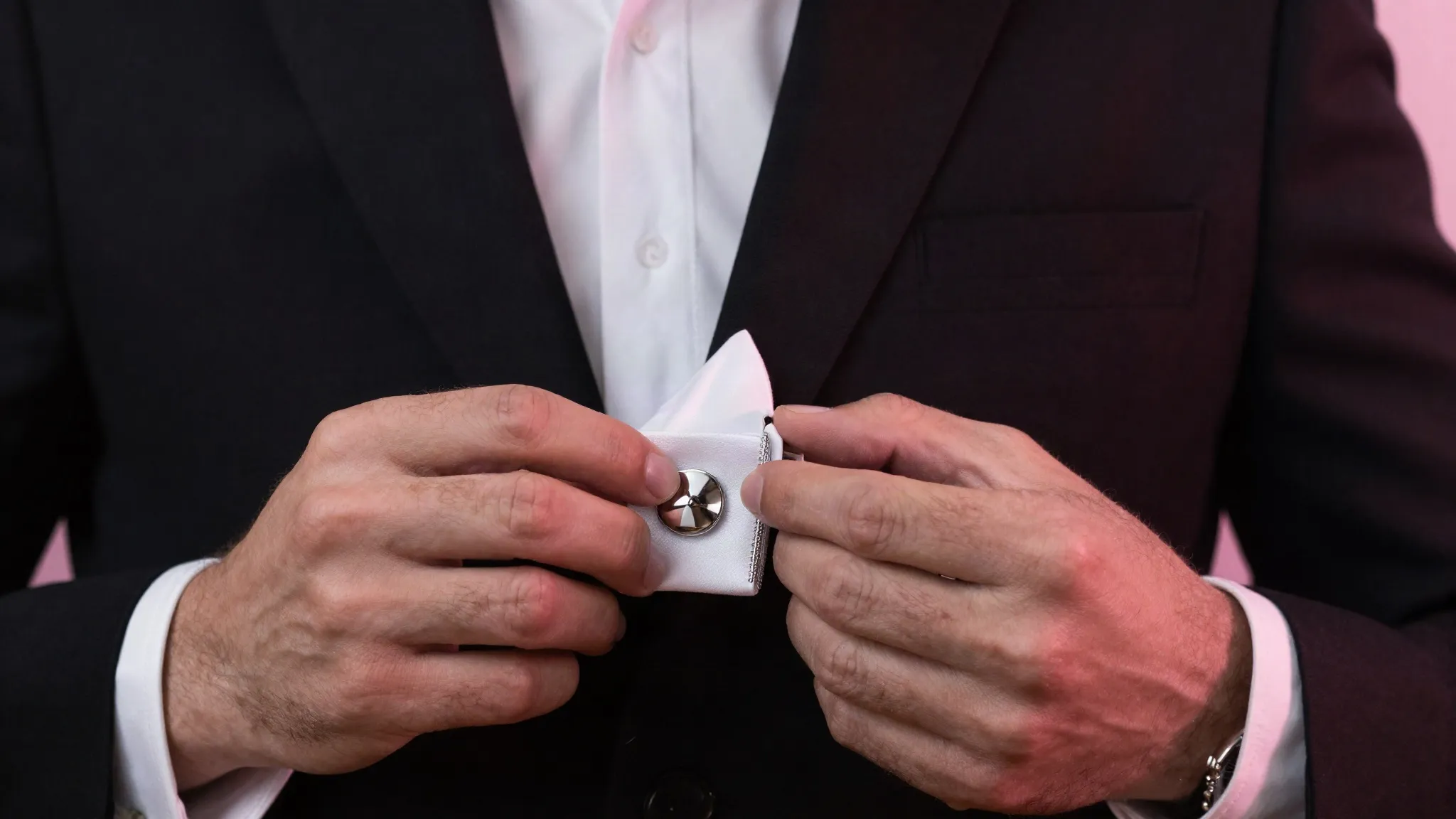 Homme ajustant le bouton de manchette d’une chemise noire sous une lumière rose
